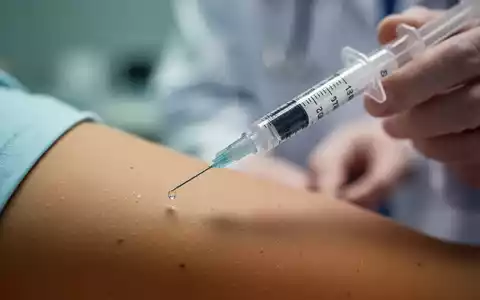 Nahaufnahme einer medizinischen Spritze mit einer FlĂŒssigkeitstropfen an der Nadelspitze, im Hintergrund unscharf ein Arm. - Foto: ĂŒber boerse-global.de