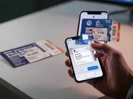Eine Hand hält ein iPhone, das die Apple Wallet App mit einem digitalen Ticket anzeigt. Im Hintergrund eine unscharfe physische Eintrittskarte. - Foto: über boerse-global.de