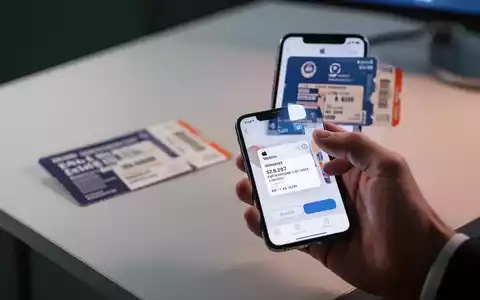 Eine Hand hält ein iPhone, das die Apple Wallet App mit einem digitalen Ticket anzeigt. Im Hintergrund eine unscharfe physische Eintrittskarte. - Foto: über boerse-global.de