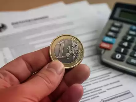 Nahaufnahme einer Hand, die eine Euro-Münze hält, mit verschwommenen Dokumenten und einem Rechner im Hintergrund. - Foto: über boerse-global.de
