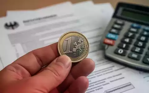 Nahaufnahme einer Hand, die eine Euro-Münze hält, mit verschwommenen Dokumenten und einem Rechner im Hintergrund. - Foto: über boerse-global.de