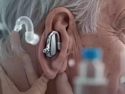 Ein modernes, diskretes Hörgerät sitzt hinter einem Ohr, im Hintergrund angedeutet eine Person oder Forschungsumgebung. - Foto: über boerse-global.de