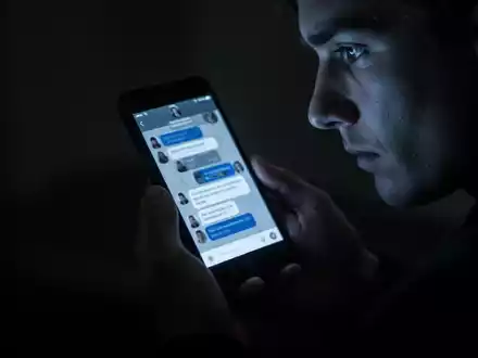 A person's hands holding a smartphone, its screen casting a dim glow on their face, reflecting a chat interface. - Foto: über boerse-global.de