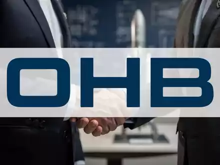 Nahaufnahme eines Händedrucks zwischen zwei Anzugträgern vor einem verschwommenen Hintergrund mit Bauplänen und einem Raketenmodell. - Foto: über boerse-global.de