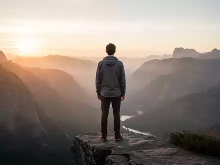 Eine Person steht auf einem Felsvorsprung und blickt auf eine weite, friedliche Berglandschaft bei Sonnenaufgang. - Foto: über boerse-global.de