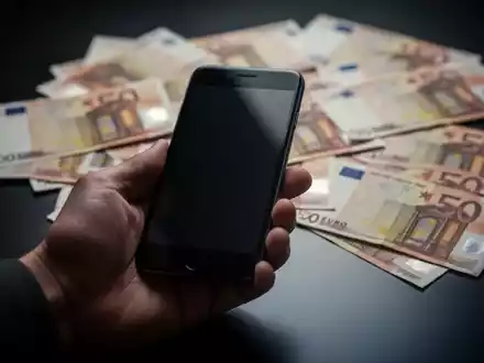 Eine Hand hält ein Smartphone, im Hintergrund unscharfe Euro-Banknoten auf einer dunklen Oberfläche. Symbolisiert Fundunterschlagung. - Foto: über boerse-global.de