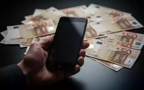 Eine Hand hält ein Smartphone, im Hintergrund unscharfe Euro-Banknoten auf einer dunklen Oberfläche. Symbolisiert Fundunterschlagung. - Foto: über boerse-global.de