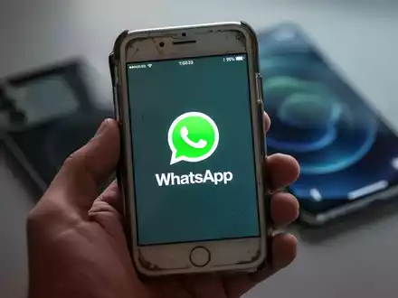 Eine Hand hĂ€lt ein Ă€lteres iPhone-Modell, auf dessen Bildschirm das WhatsApp-Symbol zu sehen ist. Im Hintergrund ist ein modernes Smartphone unscharf. - Foto: ĂŒber boerse-global.de