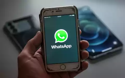 Eine Hand hält ein älteres iPhone-Modell, auf dessen Bildschirm das WhatsApp-Symbol zu sehen ist. Im Hintergrund ist ein modernes Smartphone unscharf. - Foto: über boerse-global.de