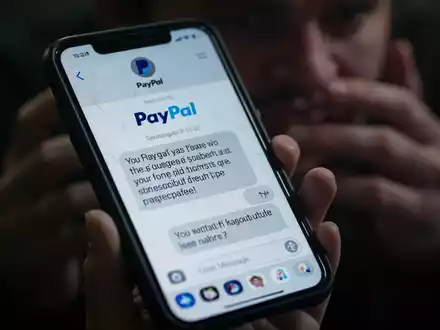 A close-up of a smartphone screen showing a fraudulent text message impersonating PayPal, held by a person with blurred hands. - Foto: über boerse-global.de
