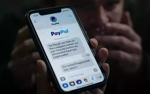 A close-up of a smartphone screen showing a fraudulent text message impersonating PayPal, held by a person with blurred hands. - Foto: über boerse-global.de