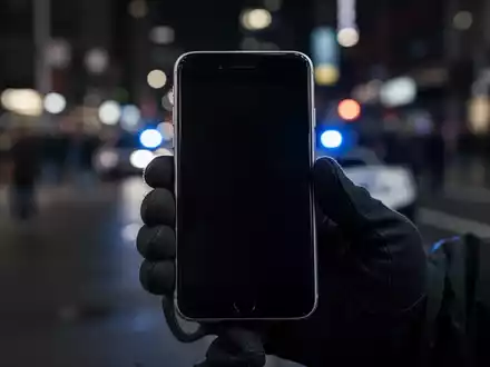A close-up of a deactivated iPhone with a dark screen, held by a gloved hand, against a blurred city street background with a police light. - Foto: über boerse-global.de