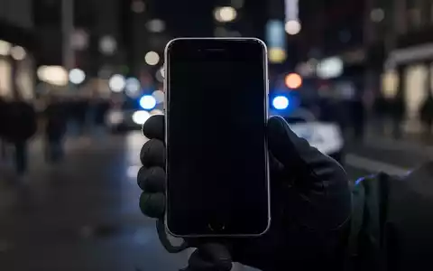 A close-up of a deactivated iPhone with a dark screen, held by a gloved hand, against a blurred city street background with a police light. - Foto: über boerse-global.de