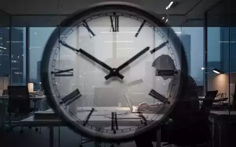 Eine Uhr mit unscharfen Zeigern, überlagert von der Silhouette einer Person, die spätabends an einem Schreibtisch arbeitet. - Foto: über boerse-global.de