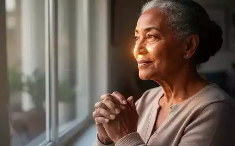 Eine nachdenkliche, ruhige Seniorin blickt aus einem Fenster, ihre Hände sanft gefaltet. - Foto: über boerse-global.de
