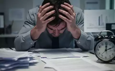 Eine Nahaufnahme der Hände einer gestressten Person, die den Kopf hält, umgeben von verschwommenen Bürodokumenten und einer Uhr. - Foto: über boerse-global.de
