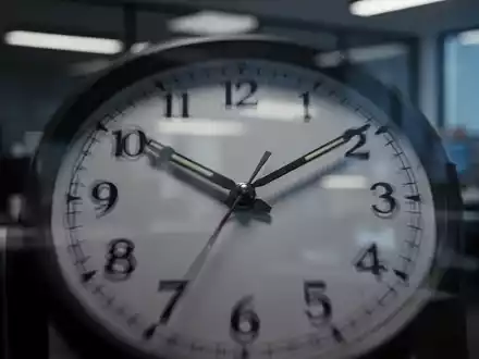 Eine Nahaufnahme einer Uhr, deren Zeiger späte Stunden anzeigen, mit einem unscharfen Büro im Hintergrund, das lange Arbeitszeiten symbolisiert. - Foto: über boerse-global.de