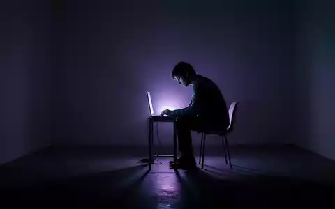 A solitary figure in silhouette sits before a glowing screen in a dark room, reflecting isolation and digital interaction. - Foto: über boerse-global.de