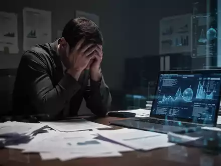 Eine Person sitzt am Schreibtisch, den Kopf in den Händen, umgeben von Dokumenten und einem Laptop, was Stress und psychische Belastung symbolisiert. - Foto: über boerse-global.de