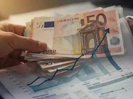 Nahaufnahme einer Hand, die Euro-Banknoten hält, vor einem Finanzdokument mit aufsteigendem Diagramm. - Foto: über boerse-global.de