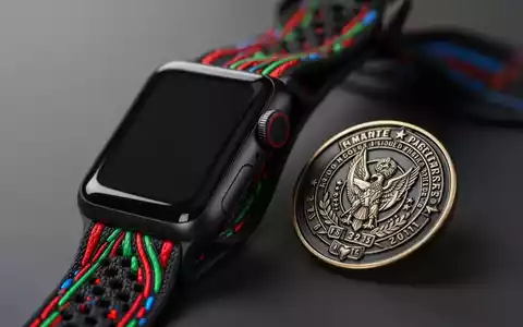 A close-up of a black Apple Watch Sport Loop band with red, green, and blue accents, next to a commemorative enamel pin. - Foto: über boerse-global.de
