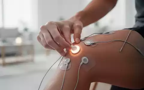 Eine Hand platziert Elektroden eines TENS-Geräts auf einem Knie, im Hintergrund ein helles, modernes Zuhause. - Foto: über boerse-global.de