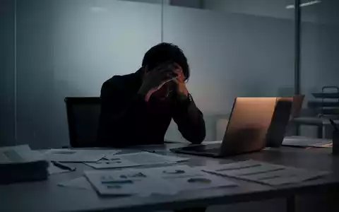 Eine einzelne, stilisierte Person sitzt gebeugt an einem Schreibtisch, den Kopf in den Händen, umgeben von verschwommenen Dokumenten. - Foto: über boerse-global.de
