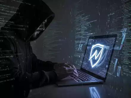 A shadowy figure in a hoodie typing on a laptop, surrounded by digital code, symbolizing cybercrime and AI scams. - Foto: über boerse-global.de
