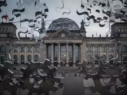 Das Reichstagsgebäude, teilweise verdeckt von abstrakten, ineinandergreifenden Zahnrädern, die Föderalismus und Finanzreform symbolisieren. - Foto: über boerse-global.de
