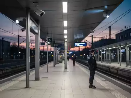 Ein sauberer, moderner Bahnhofsteig bei Dämmerung mit Sicherheitskameras und einem Polizisten im Hintergrund, der Sicherheit symbolisiert. - Foto: über boerse-global.de