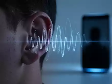 A human ear with blue light and abstract sound waves, hinting at audio manipulation, with a blurred smart device in the background. - Foto: über boerse-global.de