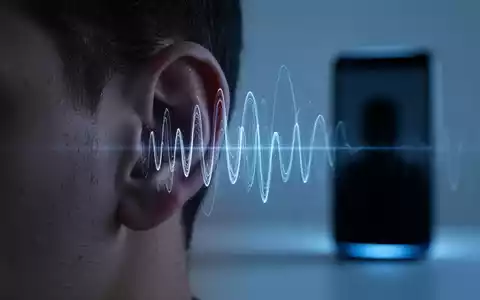 A human ear with blue light and abstract sound waves, hinting at audio manipulation, with a blurred smart device in the background. - Foto: über boerse-global.de