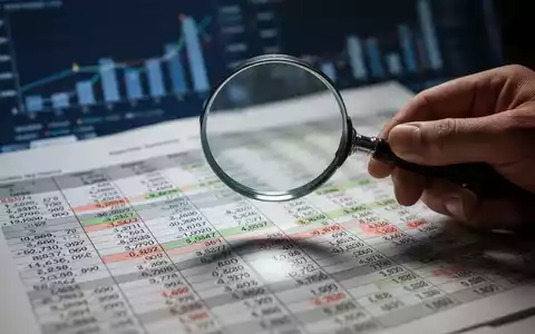 Eine Hand hält eine Lupe über ein Finanzbuch, das komplexe Einträge zeigt, im Hintergrund verschwommene Finanzdaten. - Foto: über boerse-global.de