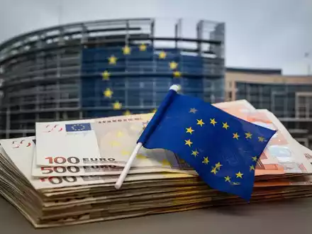 Ein Stapel Euro-Banknoten mit einer kleinen EU-Flagge davor, im Hintergrund unscharf das Europäische Parlament. - Foto: über boerse-global.de