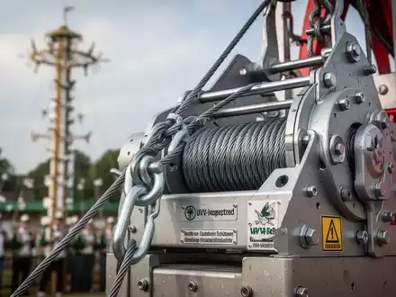 Nahaufnahme einer robusten Seilzuganlage an einer Vogelstange, im Hintergrund verschwommen ein Schützenfest. Fokus auf Sicherheitstechnik. - Foto: über boerse-global.de