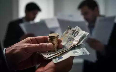 Hände halten Euromünzen und Scheine, im Hintergrund unscharf eine Person, die Dokumente prüft, symbolisiert Sozialreformen. - Foto: über boerse-global.de