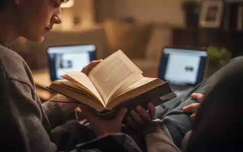 Eine Person liest konzentriert ein physisches Buch in einer ruhigen Umgebung, mit unscharfen digitalen Geräten im Hintergrund. - Foto: über boerse-global.de