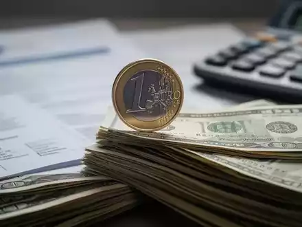 Eine einzelne Euromünze balanciert auf einem Stapel Geldscheine, im Hintergrund unscharfe Dokumente und ein Taschenrechner. - Foto: über boerse-global.de