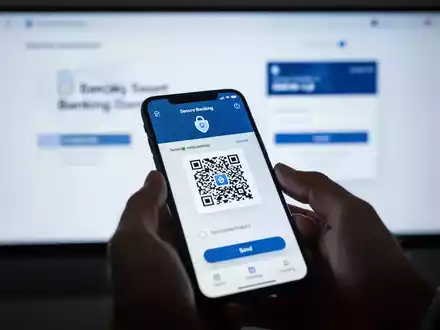 A hand holds a smartphone displaying a banking app with a QR code, with a blurred computer screen in the background. - Foto: über boerse-global.de