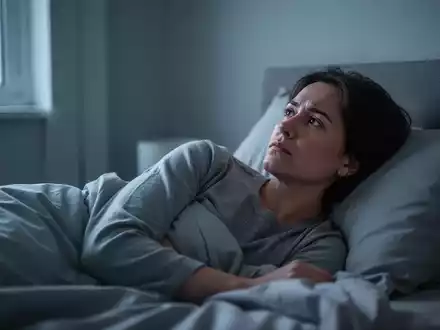 Eine Person liegt wach und nachdenklich im Bett, umgeben von kühlem Umgebungslicht, das durch ein Fenster fällt. - Foto: über boerse-global.de