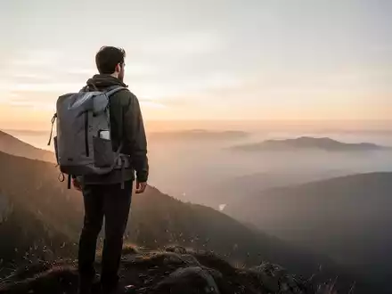 Ein einsamer Wanderer mit minimalistischem Rucksack auf einem Bergrücken bei Sonnenaufgang, der über eine weite, neblige Landschaft blickt. - Foto: über boerse-global.de