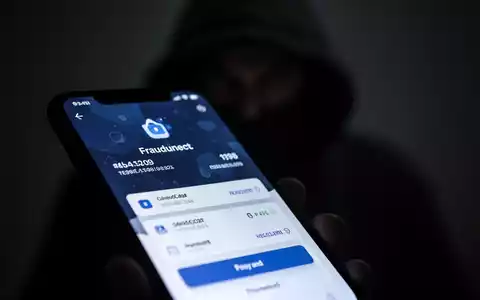 Ein Smartphone-Bildschirm zeigt eine betrügerische Zahlungs-App, im Hintergrund eine unscharfe, schattenhafte Hacker-Figur. - Foto: über boerse-global.de
