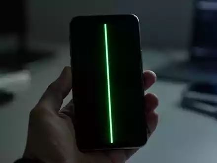 A smartphone screen displaying a prominent, glowing green vertical line, held by a hand in a dimly lit environment. - Foto: über boerse-global.de