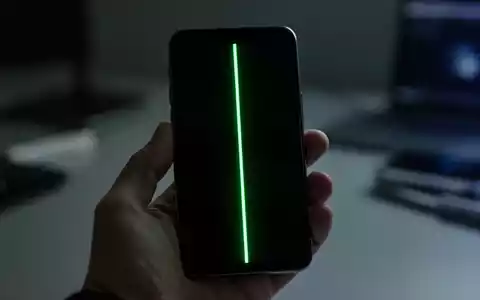 A smartphone screen displaying a prominent, glowing green vertical line, held by a hand in a dimly lit environment. - Foto: über boerse-global.de