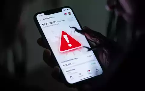 Nahaufnahme eines Smartphone-Bildschirms mit einer Banking-App, die ein rotes Warnsymbol zeigt, gehalten von einer Hand im Schatten. - Foto: über boerse-global.de