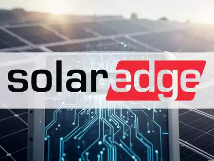 Nahaufnahme eines modernen Solar-Wechselrichters mit sichtbarer Schaltung vor einem unscharfen, sonnenbeschienenen Solarmodulfeld. - Foto: über boerse-global.de