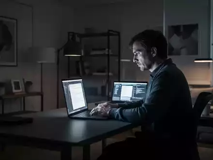Eine Person arbeitet alleine an einem Laptop in einem modernen, schwach beleuchteten Heimbüro, symbolisiert Isolation und Technologieeinsatz. - Foto: über boerse-global.de