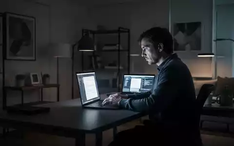 Eine Person arbeitet alleine an einem Laptop in einem modernen, schwach beleuchteten Heimbüro, symbolisiert Isolation und Technologieeinsatz. - Foto: über boerse-global.de