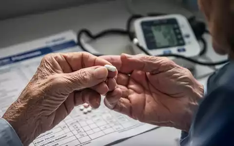 Nahaufnahme der Hand einer älteren Person, die eine Pille hält, mit einem unscharfen Hintergrund, der ein medizinisches Diagramm oder einen Blutdruckmesser zeigt. - Foto: über boerse-global.de