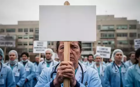 Ein Nahaufnahme der Hände eines Krankenhausmitarbeiters, der ein leeres Streikschild hält, im Hintergrund unscharf weitere Streikende und ein Krankenhaus. - Foto: über boerse-global.de